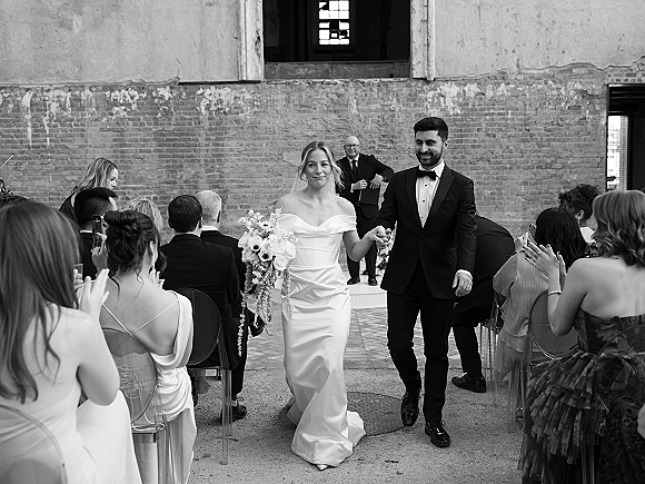 Wedding recessional as newlyweds walk down aisle holding hands, bride with cascading bouquet in off-shoulder gown, guests clapping in brick courtyard