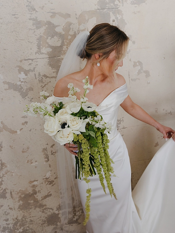 Bridal portrait of a bride in an off the shoulder wedding dress with veil and pearl drops, holding a white and green bouquet by a plaster wall