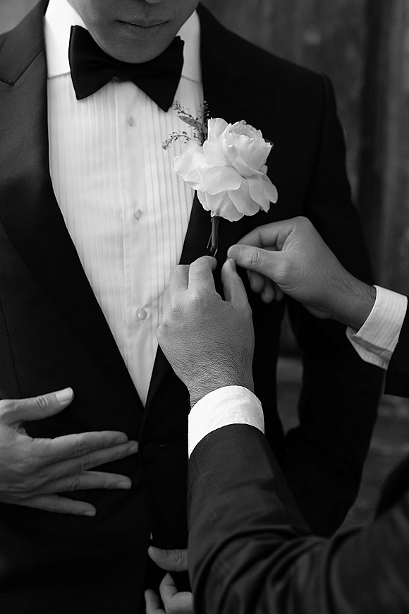 Groom getting ready as he pins a white rose boutonniere on his black tuxedo lapel, bow tie and pleated shirt in a blurred interior