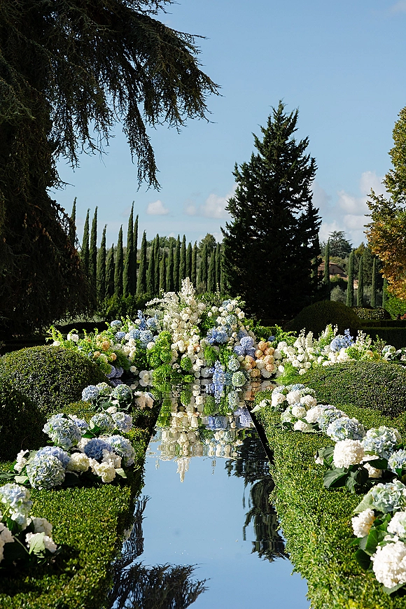 Ceremony aisle decor with mirrored aisle runner, lined by hydrangea and white pastel florals reflecting water in a formal garden setting