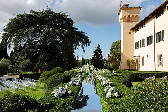 Outdoor ceremony setup with mirrored wedding aisle, clear acrylic chairs and hydrangea floral arrangements on a manicured villa lawn under blue sky
