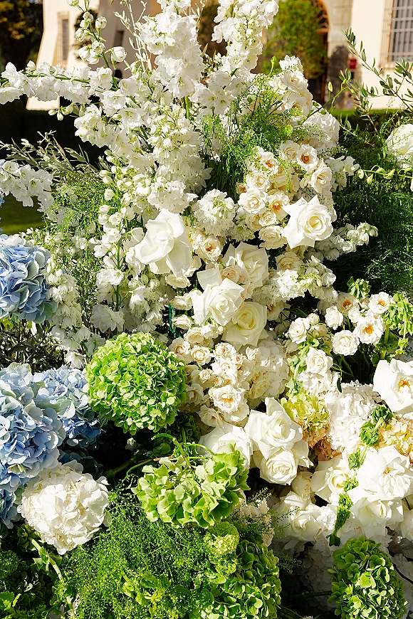 Wedding floral arrangement of white rose wedding flowers with hydrangea, delphinium, and greenery on a sunlit garden lawn by a stone facade