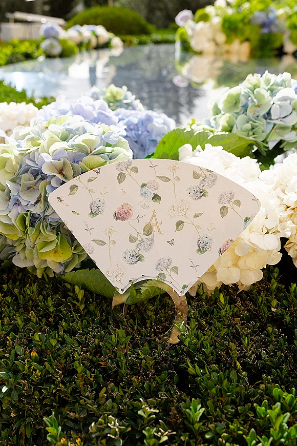 Wedding program displayed on a clear acrylic stand with a floral illustrated program fan and hydrangea blooms by a garden reflecting pool
