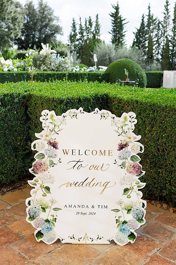 Wedding welcome sign with gold lettering and hydrangea floral illustration on a scalloped board, displayed on an easel in a garden patio setting