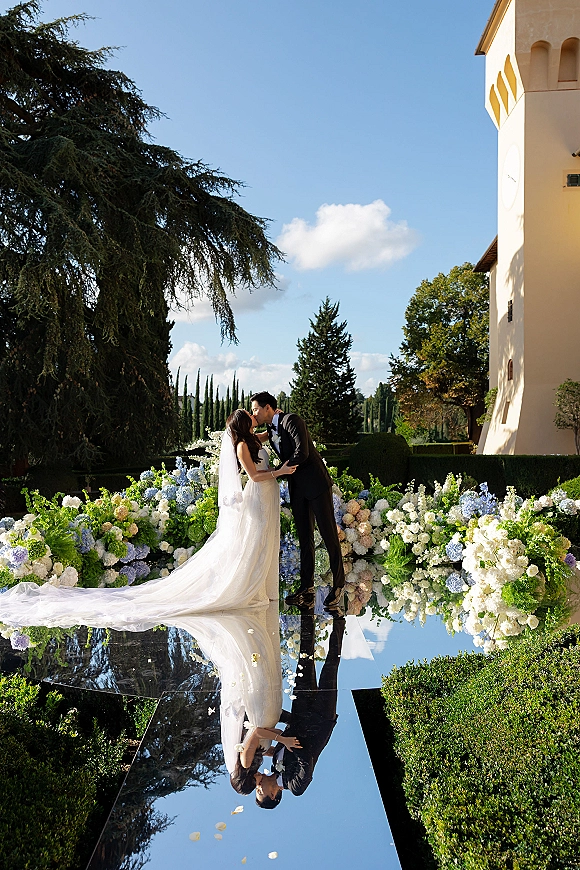 Wedding kiss on a mirrored aisle, bride and groom kissing in veil and tuxedo amid hydrangea florals in a hedge garden by a villa