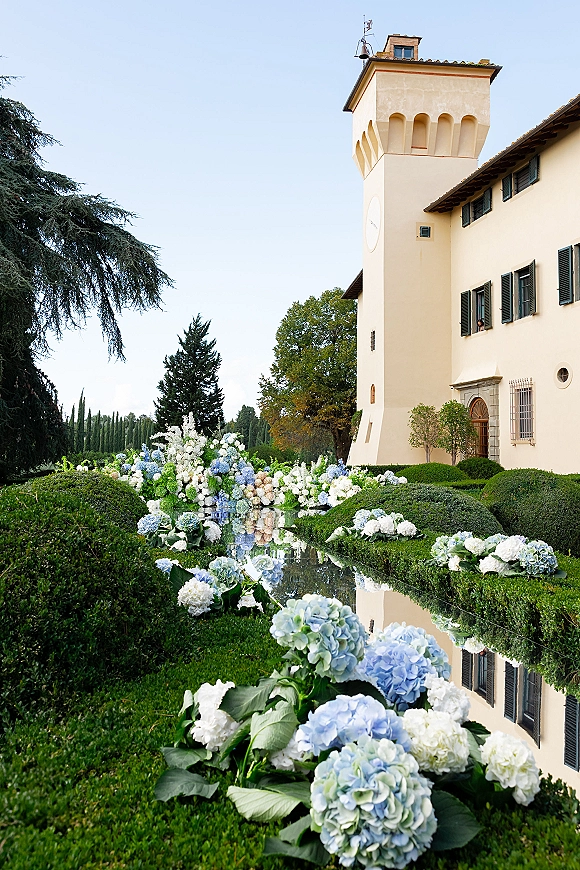 Outdoor wedding florals with hydrangea wedding flowers lining a reflecting pool, framed by manicured hedges and a villa tower backdrop