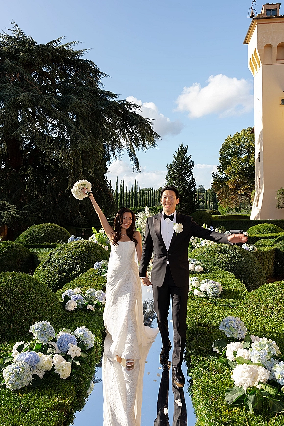 Newlywed couple portrait walking a mirror wedding aisle, bride raising bouquet, groom in black tuxedo, hedged villa garden backdrop