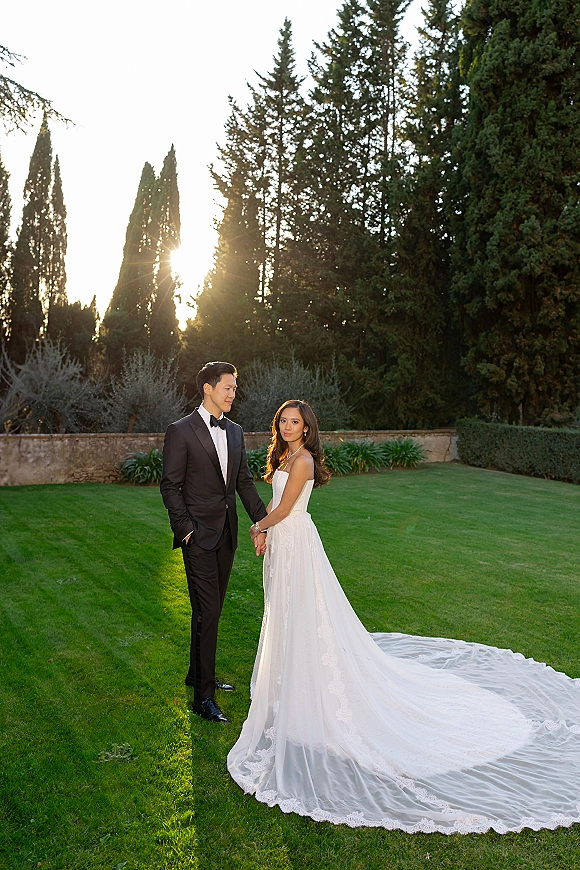 Couple portrait of bride and groom holding hands, her strapless dress with lace train and his tuxedo, on a tree-lined lawn with sun flare