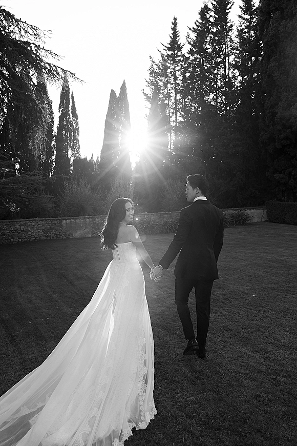 Couple portrait of bride and groom walking away hand in hand, bride looking back as her long lace train trails on a lawn by trees and stone wall with sun flare