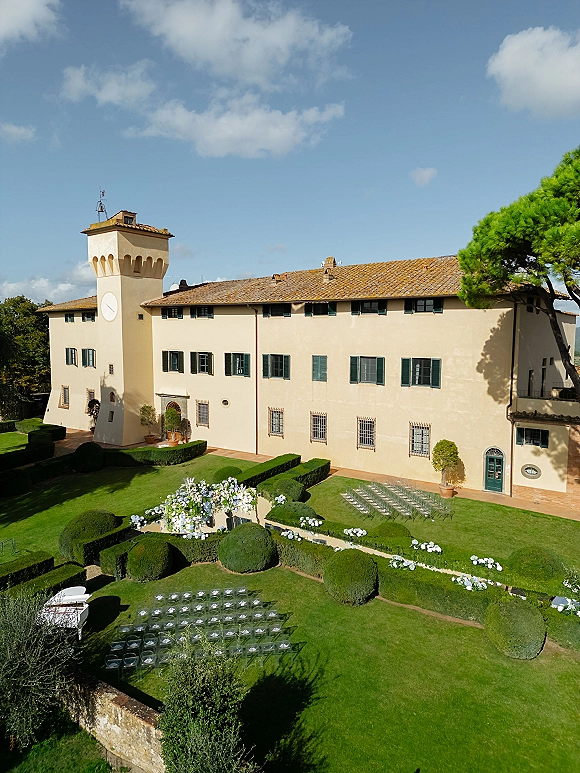 Outdoor ceremony setup with garden wedding ceremony arch of white and blue flowers, clear chairs on courtyard lawn before villa clock tower