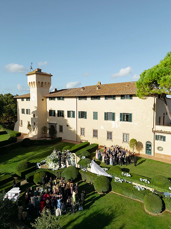 Outdoor wedding ceremony in a manicured garden with a white aisle runner, floral arrangements, and a bride’s long train by a historic villa clock tower