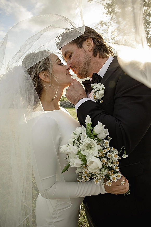 Wedding kiss portrait of bride and groom kiss under a flowing veil, bride in long sleeve gown holding white daisy bouquet in a sunlit field