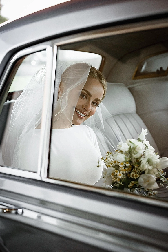 Bridal portrait of a bride in car, smiling behind her veil in a leather backseat, holding a white and green bouquet by the window