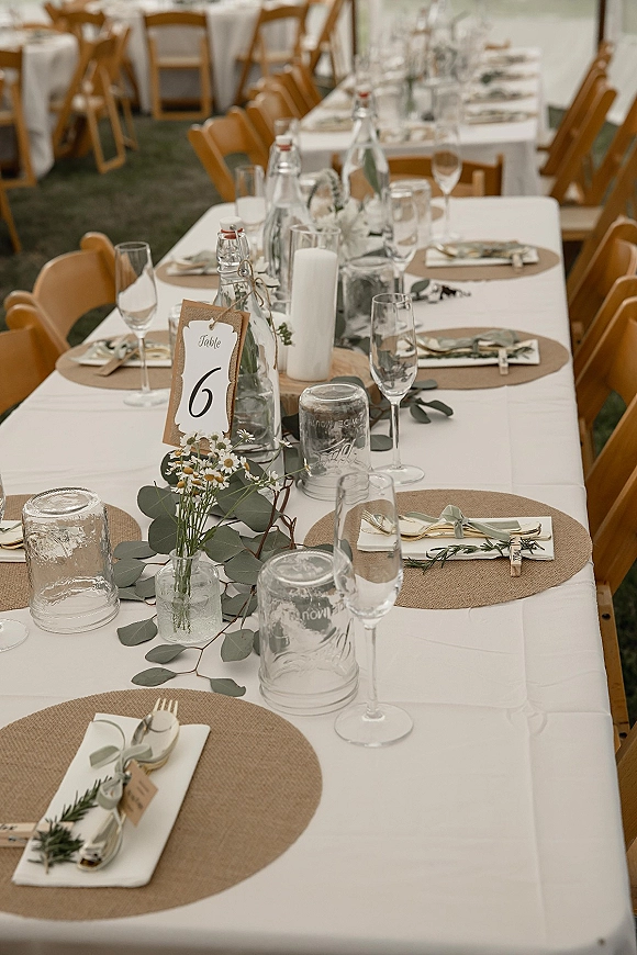 Reception tablescape with rustic wedding table setting: white linen, burlap placemats, daisies, eucalyptus greenery, candles on lawn banquet tables