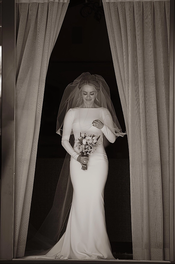 Bridal portrait of a bride holding bouquet with a veil over her face, looking down in a long sleeve dress at a dark doorway with curtains