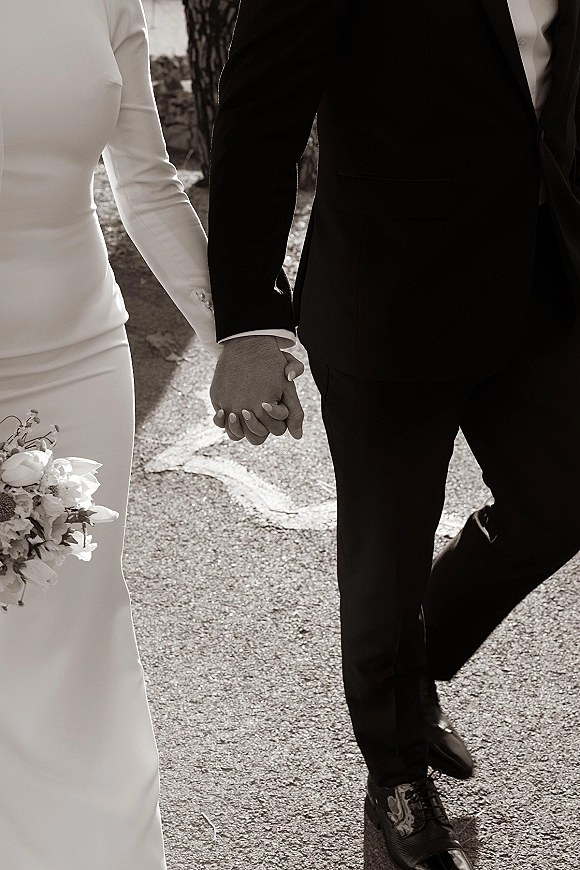 Couple holding hands, bride and groom walking on a tree-lined paved road, showing wedding rings, bouquet, and long sleeve dress