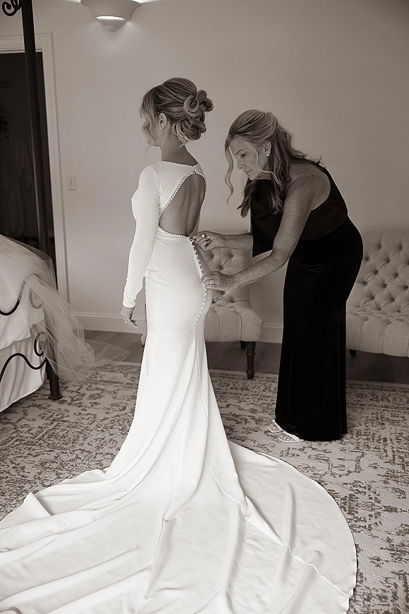 Bride getting ready as a mother buttons her pearl-trim open-back wedding dress with long sleeves and train in a cozy bedroom setting