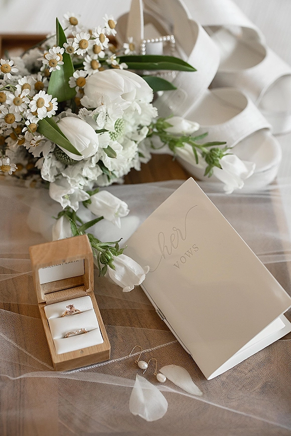 Wedding detail flatlay with wedding ring box, white tulips and chamomile bouquet, vow book, pearl earrings, veil, and shoes on a wood table