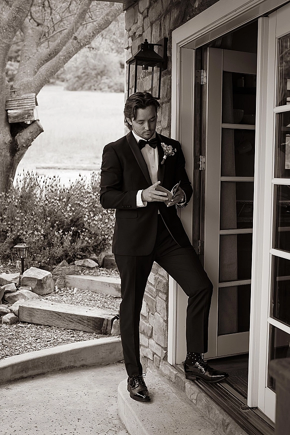 Groom portrait in a black tuxedo with bow tie and boutonniere, standing by a stone doorway on an outdoor patio with shrubs