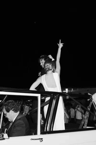 Wedding exit as the couple in getaway car, bride in low back dress waving from a vintage convertible under night sky, guests cheering