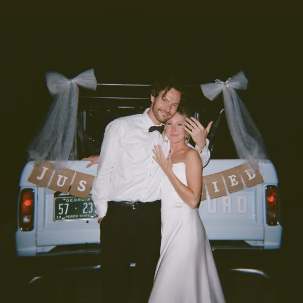 Couple portrait of newlyweds holding up rings beside a wedding getaway car with just married banner on a vintage pickup truck at night