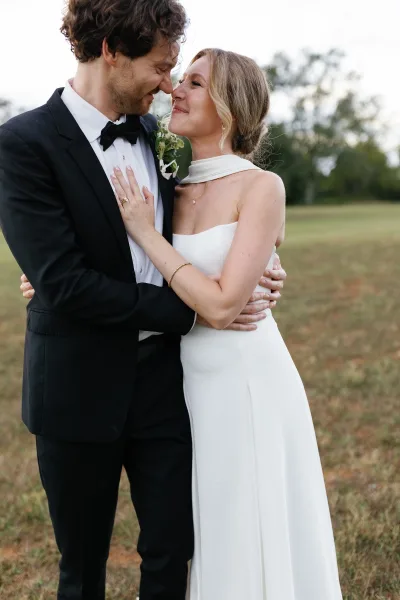 Couple portrait of bride and groom embrace, forehead touch as she holds his lapel, showing rings in a field with trees and sky