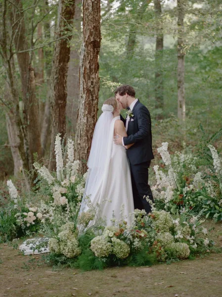 Wedding kiss as bride and groom kissing on a woodland dirt path, her veil trailing beside white ground florals and greenery