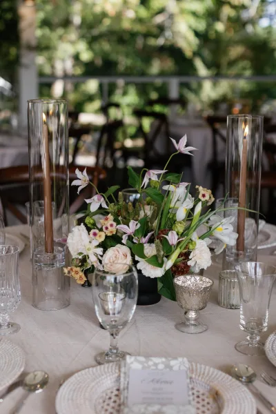 Reception tablescape with a wedding table centerpiece of blush and white blooms, taper candles, silver goblets, and rattan chargers on an outdoor patio
