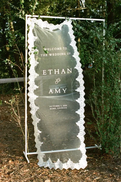 Wedding welcome sign on a white metal frame, an acrylic wedding welcome sign with lace trim, set along a wooded path by a fence