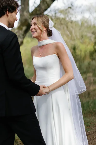 Ceremony moment of bride and groom holding hands as they exchange vows, her veil and lace headpiece framed by daylight trees and greenery