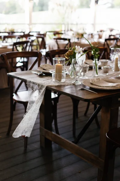Reception tablescape with wedding farm table decor featuring a lace runner, taper candles, glass goblets, woven chargers, and floral centerpieces on a covered patio