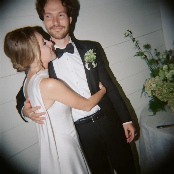 Couple portrait of bride and groom embrace, her sleeveless gown and his black tuxedo, posed by a reception table on white shiplap wall