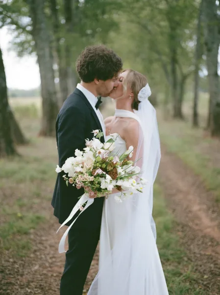 Wedding kiss portrait of bride and groom kissing, bride holding bouquet with ribbon and veil bow on a dirt path among trees