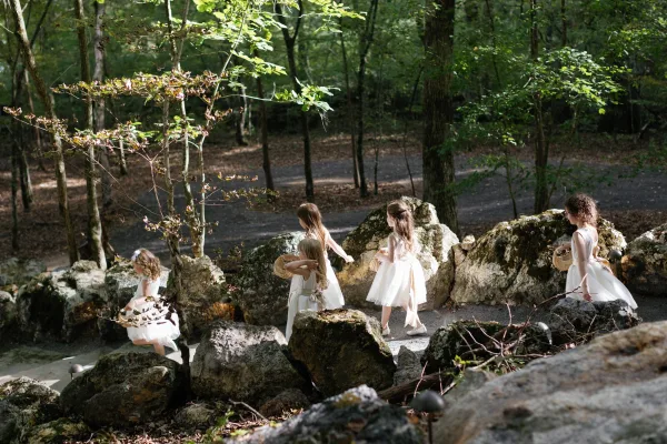 Flower girl moment as flower girls walking in tulle dresses with basket purses and hair bows along a forest gravel path in dappled sunlight