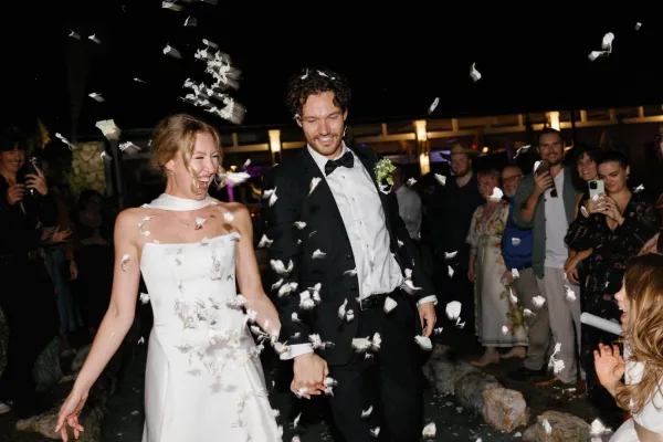 Wedding exit as bride and groom walk hand in hand through petal confetti, guests filming on phones under warm lights outside at night