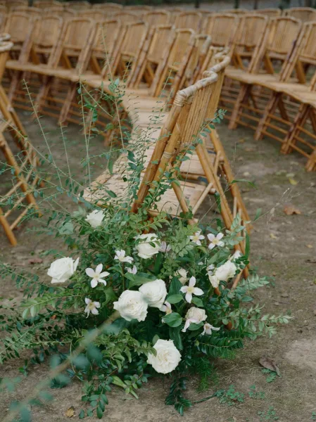 Ceremony aisle decor with wedding aisle flowers of white roses and greenery along dirt ground, flanked by rows of wood folding chairs