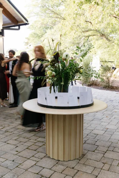 Wedding seating chart escort card display on a round pedestal table with white florals and greenery, set on patio pavers with guests nearby