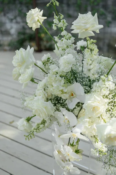Wedding floral arrangement with a white wedding centerpiece of roses, orchids, hydrangea, baby's breath, and greenery on a white bench by a stone wall