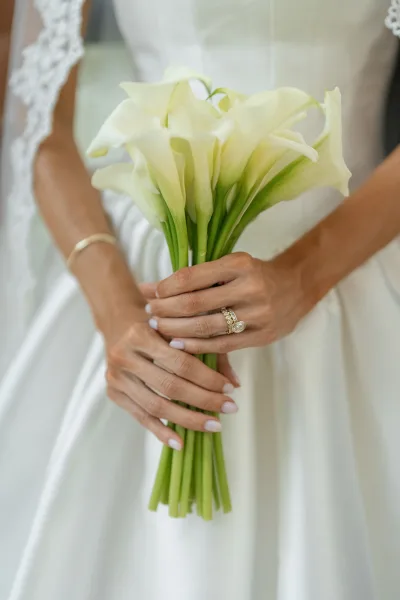 Calla lily bouquet held in bride’s hand with pale pink manicure, gold bracelet, and wedding ring set against white dress fabric