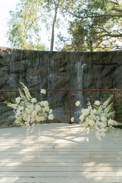Wedding altar decor with asymmetrical altar flowers on clear acrylic pedestals, white roses, orchids, and greenery before a stone waterfall wall.