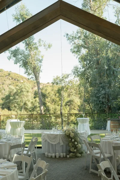 Reception tablescape with white wedding reception tables set with candles, glassware, and floral centerpieces inside a glass pavilion with hillside views