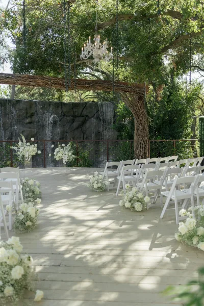 Ceremony setup with white folding chairs facing a wooden arch and chandelier, framed by white flowers and greenery under trees by a stone wall