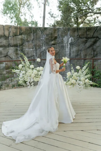 Bridal portrait of a bride in a wedding dress with train, holding a white calla lily bouquet beside a stone waterfall wall outdoors