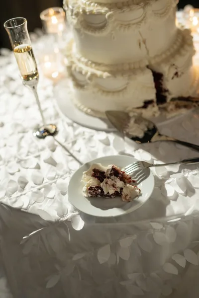 Wedding cake with white tiered wedding cake piping beside a slice on dessert plate, silver server, champagne flute, and votive candles against bokeh lights