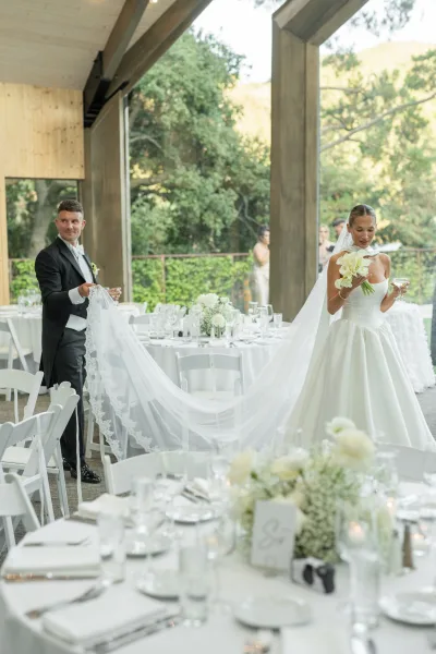 Wedding reception moment as bride holding bouquet and champagne coupe while groom lifts her long lace veil on a covered patio with white tables and candles