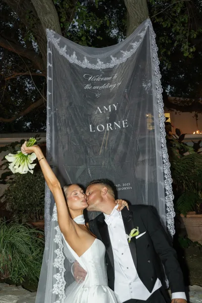 Wedding kiss portrait of bride and groom kissing under a lace veil, her calla lily bouquet raised beside a welcome sign in a garden courtyard