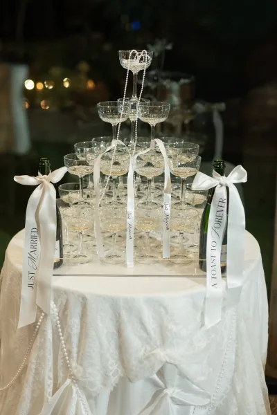 Champagne tower setup with a champagne coupe tower on a lace-draped table, white ribbon bows and pearl garland in a dark reception space