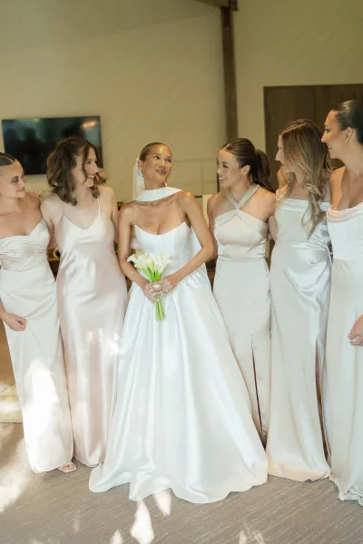 Bride with bridesmaids in champagne bridesmaid dresses gathered indoors, bride in veil and strapless gown holding a white calla lily bouquet