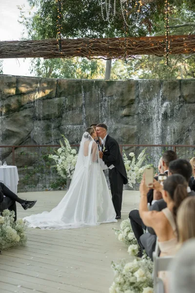 Wedding kiss as bride in cathedral veil and long train leans into groom in tux before a stone waterfall wall and string lights