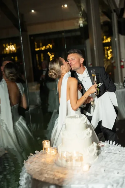Wedding cake cutting as bride and groom kiss beside a tiered white cake with champagne flutes and candles, string lights reflected on glass wall